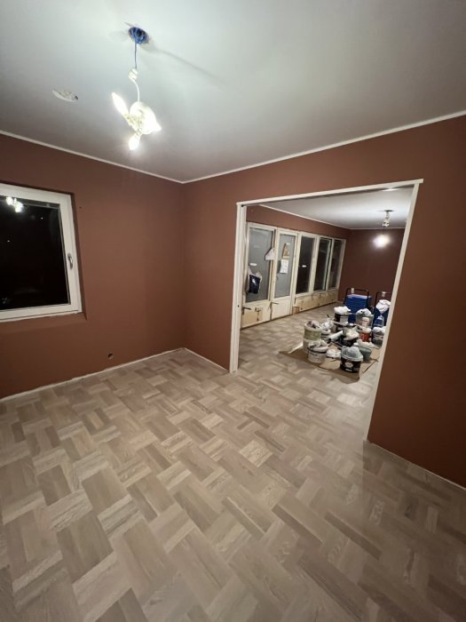 Ett tomt rum med brunmålade väggar och nytt parkettgolv. En lampa i taket och byggmaterial staplade längst väggen.