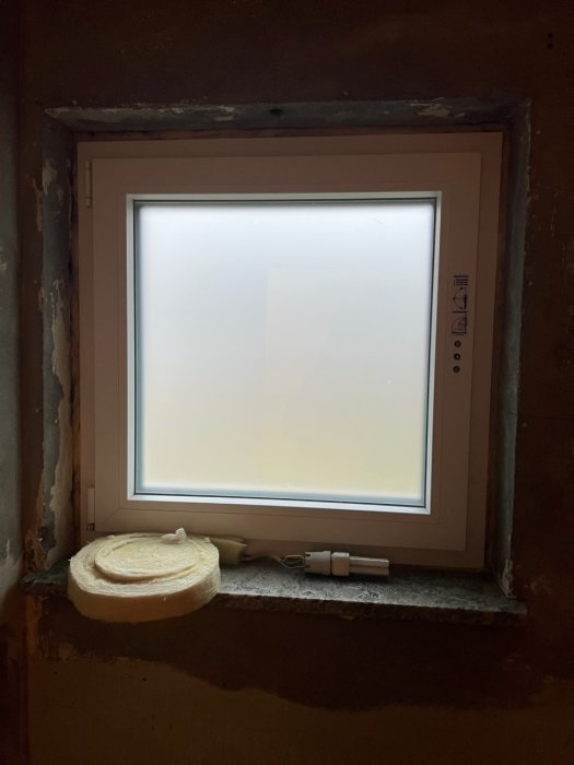 Ett suddigt fönster inbäddat i en ojämn murad vägg med synlig isolering och elektriska komponenter nära fönsterbrädan.