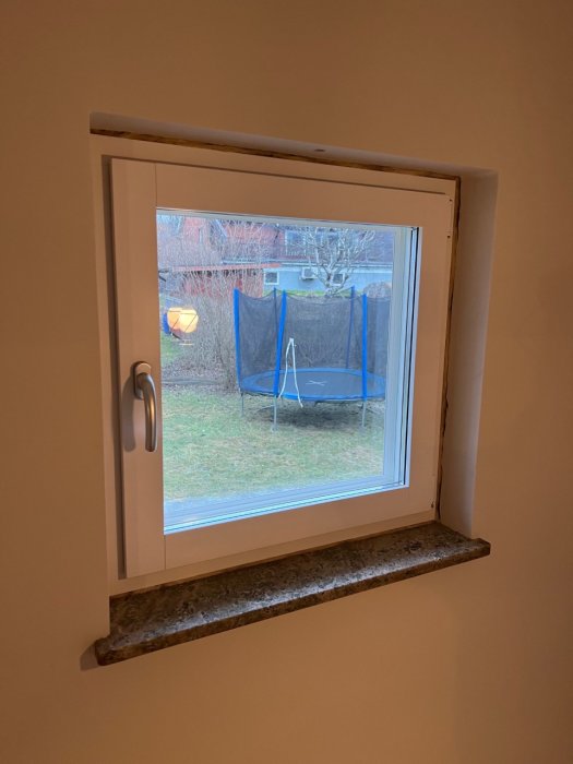 Ett fönster med utsikt över en trädgård, trampolin synlig, grått stenfönsterbräde, delvis öppet fönster.