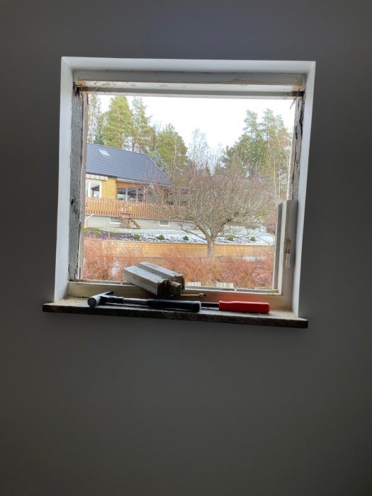 Ett fönster med verktyg, utsikt över träd, hus och snöfläckar. Slitet fönsterbräde, inomhusmiljö.