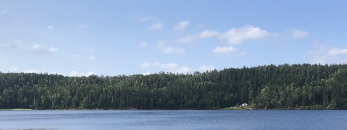 Ett lugnt sjölandskap med skogsbeklädda berg och klarblå himmel. En liten byggnad vid sjökanten.