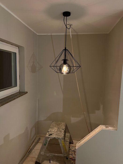 Ett rum under renovering med tänd lampa, stege, kala väggar och en fönsterkarm.