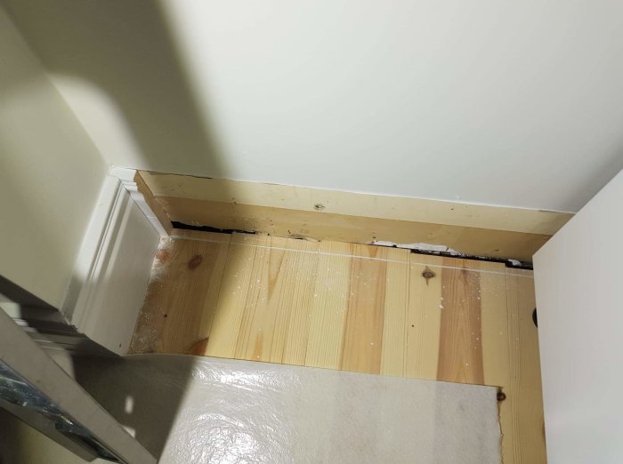 Hörn av ett rum med trasig golvlist och synliga spikar, behöver reparation eller renovering.