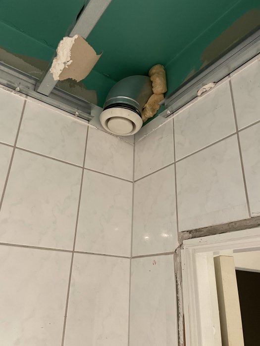 Luftventil i hörn, skadat takgips, frigolitisolering synlig, kaklade vita väggar, grön vägg ovanför. Behov av reparation.