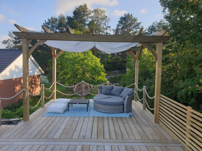 Trädäck med pergola, hängstol och soffmöbler, omgiven av natur och repstaket.