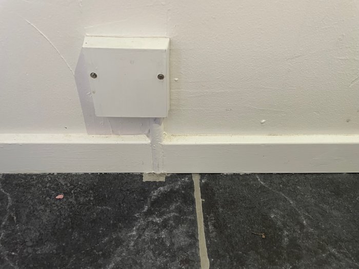 Vit vägg, elektrisk kabel, grått golv, liten vit låda, kablageuttag, spricka i väggen, lister, nedsmutsat golv.