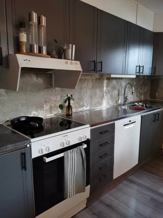 Modernt kök med mörka skåp, vita vitvaror, rostfri diskho och rå betongvägg.