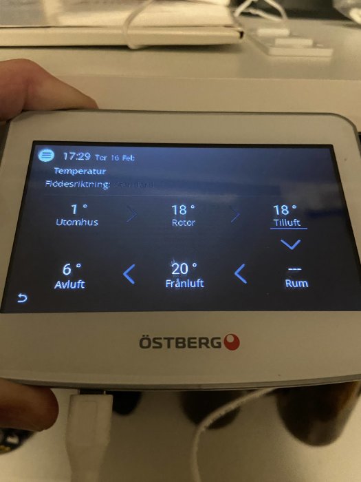 En hand håller en Östberg termostat visande tider, datum, och olika temperaturinställningar på svenska.