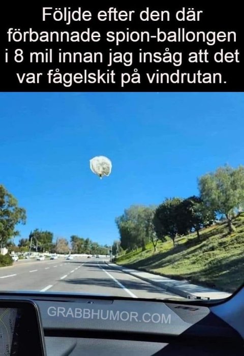 Bilvy med väg och träd, fågelspillning på vindruta ser ut som ballong i himlen. Humoristisk text.