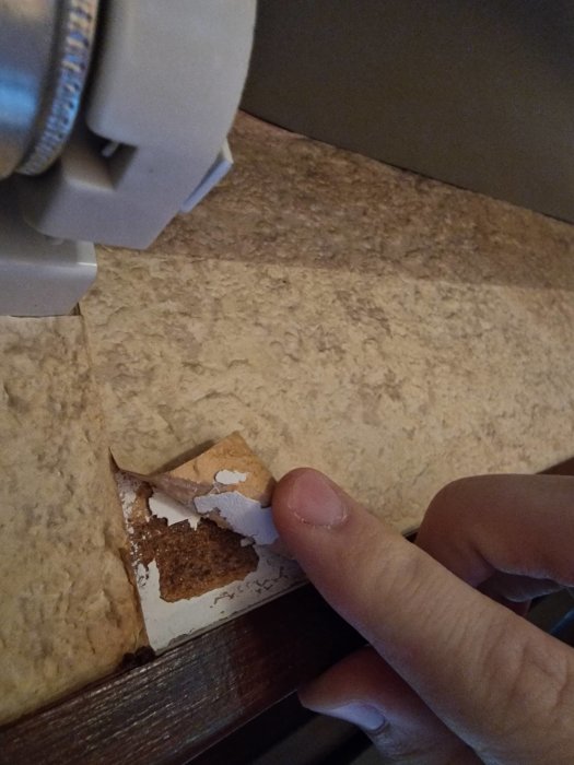 Finger drar bort lager av murket material, avslöjar underliggande brunt lager, nära en ljusarmatur i bakgrunden.