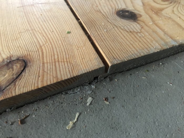 Trägolvplankor med avstånd emellan, visande nod och sågmärken, på grått betongunderlag. Lite smuts och skräp synligt.