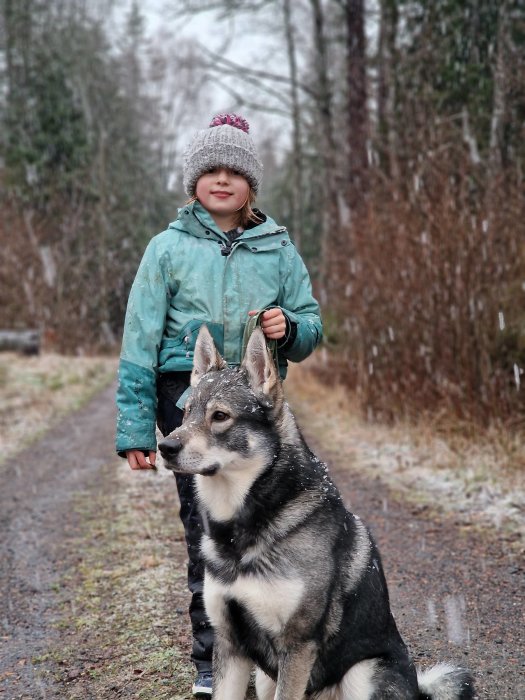 Barn i mössa och jacka med en hund på en snöig skogsväg.