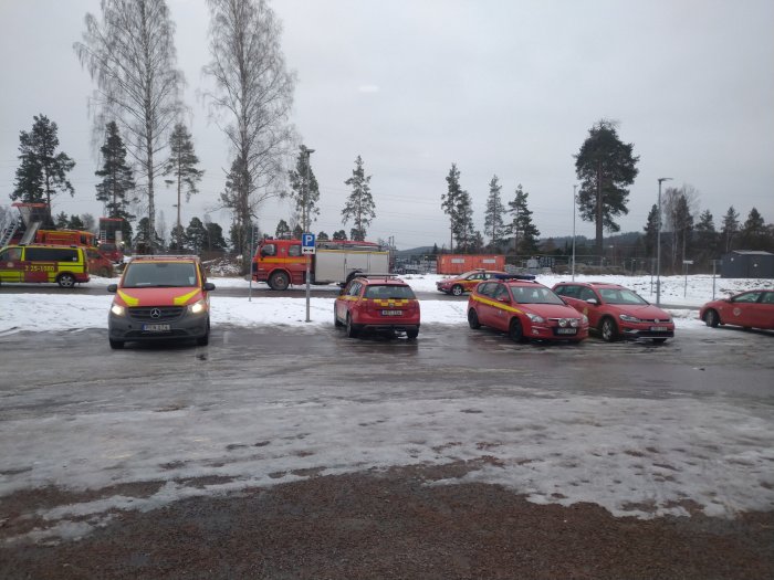 Parkeringsplats med bilar, snöfläckar, brandbilar i bakgrunden, gråmulet väder.