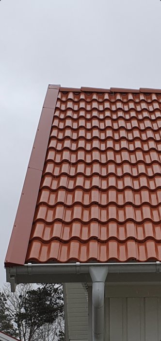Del av hus med tegelröd takpannor, snedtak, vit stupränna och fasaddetaljer, grå himmel.