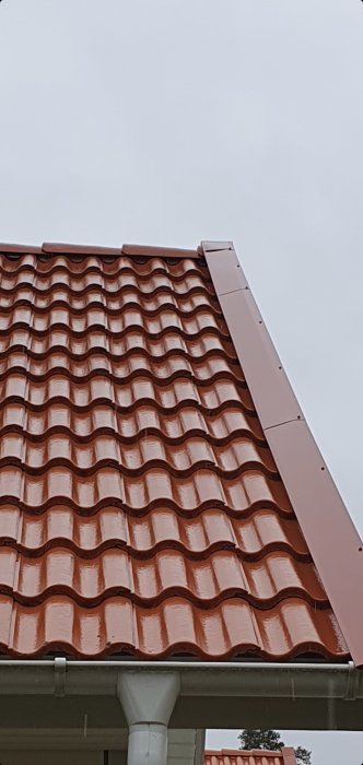 Tak med röda tegelpannor, vit stupränna, grå himmel, hörn på husfasad, byggnadsdetaljer, nordisk arkitektur.