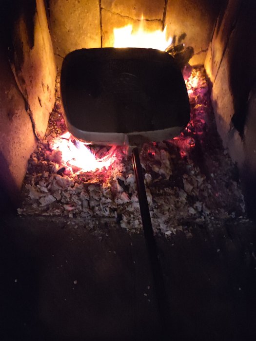 Stekpanna värms i öppen eld i eldstad, glödande kol syns. Matlagningsförberedelser, mysig atmosfär, rustikt.