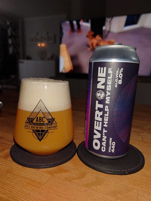 Ölglas med skum på underlägg bredvid ölburk, ABC-logotyp, inomhus och TV i bakgrunden.