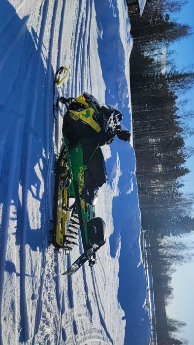 Snöskoter ligger på sidan i snön, klart väder, blå himmel, skogsbakgrund.