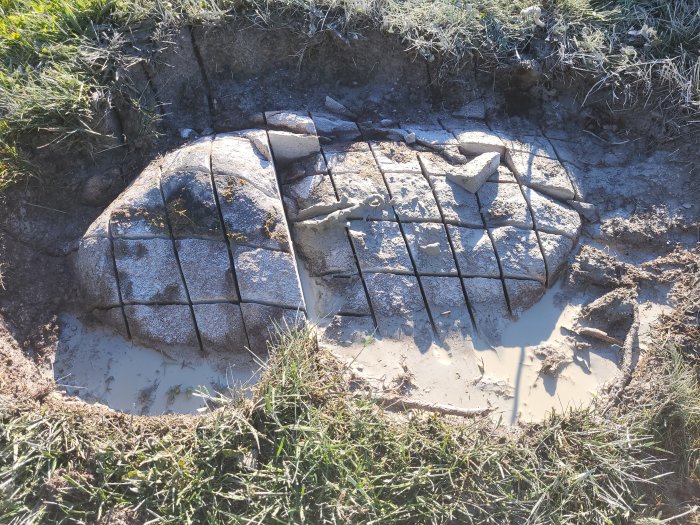 Sönderdelad betongväg med armeringsjärn i en pöl av vatten och lera omgiven av gräs.