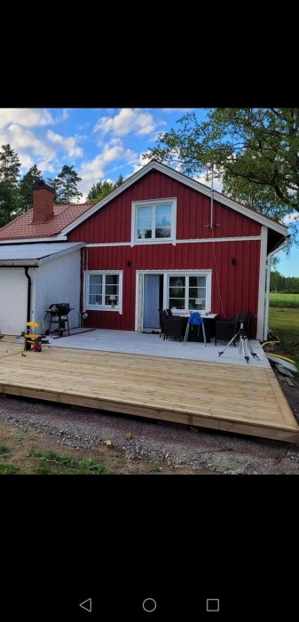 Rött hus med vit knut, nybyggd träveranda och grill, klar himmel, gröna träd i bakgrunden.