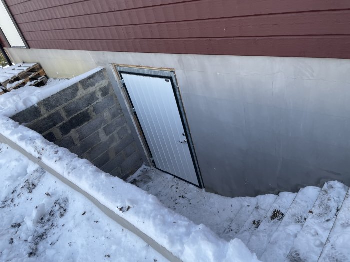 Källardörr i betongvägg, trappa med snö, röd byggnad, vinterdag.