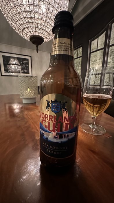 En ölflaska och ett tomt glas på ett bord, ljuskrona i bakgrunden, mysig atmosfär.