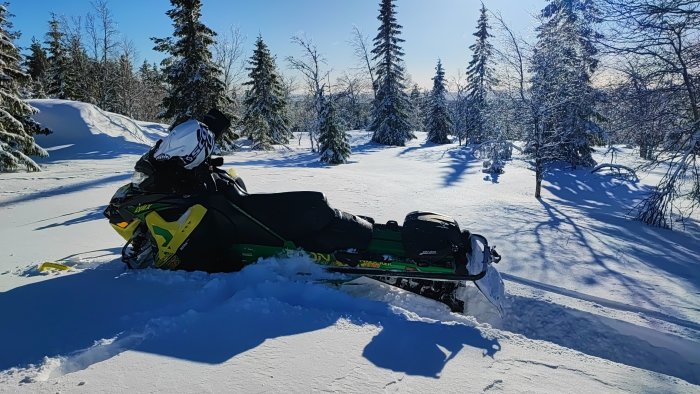 Snöskoter och förare i vintervitt landskap med snötäckta träd och klarblå himmel.