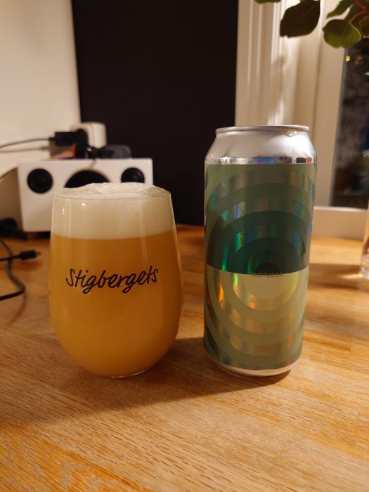 Ett ölglas med skum på ett bord, bredvid en ölburk med etikett. Bakgrund: rumsinredning, belysning.