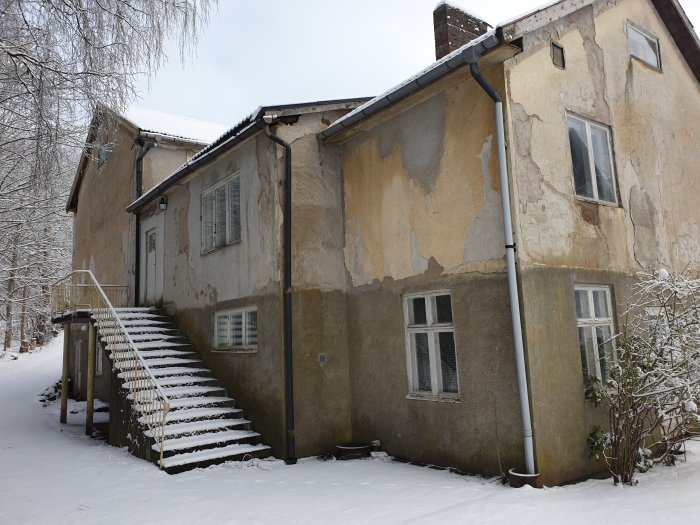 Äldre hus med sliten fasad, snöbelagd trappa, vinterscen, kala träd, vitt tak, frostigt väder.