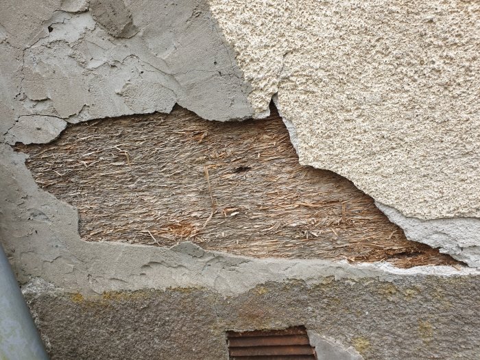 Slitet ytterskikt på vägg avslöjar underliggande stråmaterial. Skadad puts. Struktur- och texturskillnader synliga.