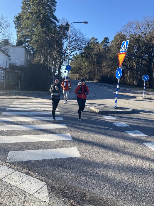 Tre personer springer över ett övergångsställe på en solig dag, omgivna av gatuskyltar och träd.