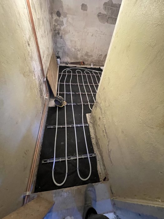 Nedgång till en trång källare med golvvarmerör installerade, orenoverade väggar, arbetsområde, bygghjälm synlig.