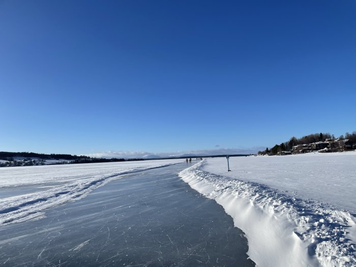 Klarblå himmel, frusen sjö, folk åker skridskor, snödriva, bostadshus vid strandkanten, solig vinterdag.