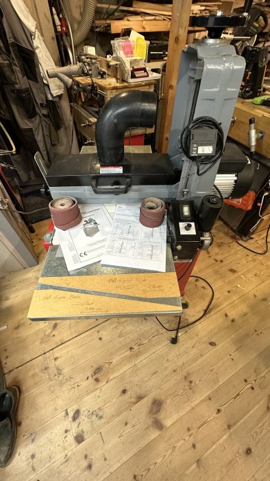 Verkstad med slipsmaskin, dammutsug, arbetsskiss, trästycken och elektriska verktyg på ett trägolv.