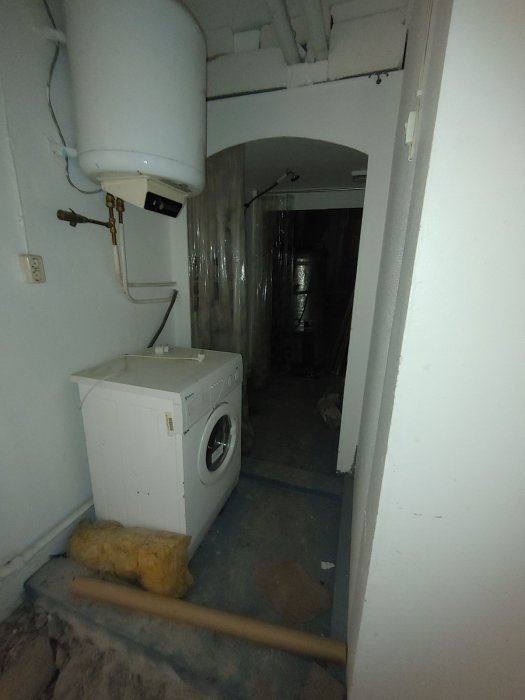 Förfallen källare med tvättmaskin, varmvattenberedare, isolering, rör, och oavslutad renovering.