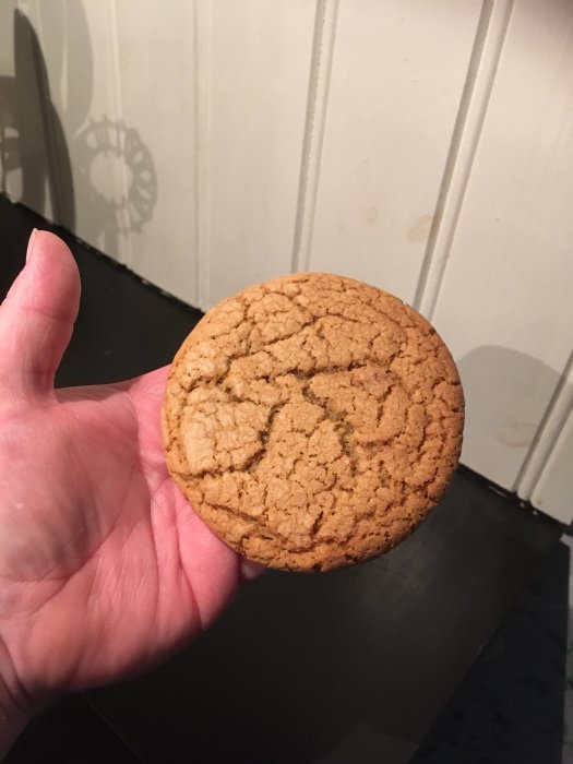 En hand håller en stor, gyllenbrun kaka framför en vit vägg.