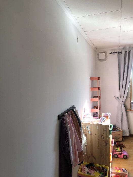 Ett rum med leksaker, kläder på krokar, en vit vägg och gardiner vid fönstret.