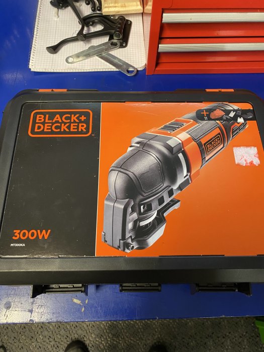 Black & Decker verktygslåda, troligtvis innehållande en slipmaskin, på ett verkstadsbord med verktyg och lådor i bakgrunden.