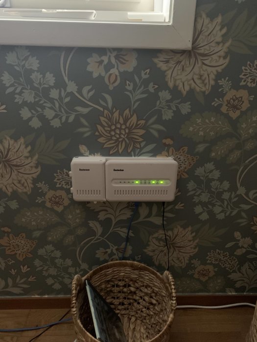 Två white routers med lysande lampor monterade på vägg med blommigt mönster, kablar nertill, korg i förgrunden.