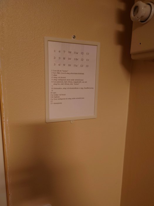 Upphängd tavla med text, sannolikt instruktioner eller schema, på en gul vägg vid en strömbrytare.