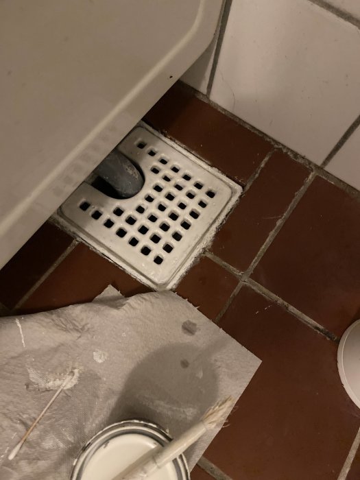 Golvbrunn bredvid toalett, pappershandduk, pensel, färgburk, vita fläckar på brunt klinkergolv.