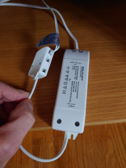 En hand håller en nätadapter med flera USB-portar och anslutna kablar mot ett träbord.