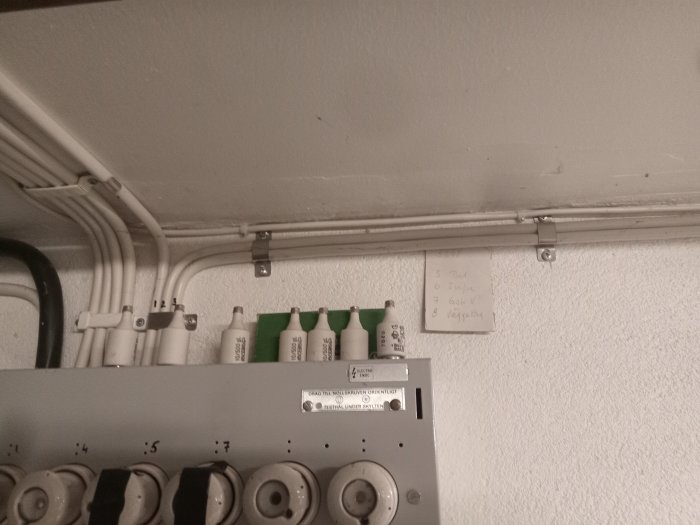 Elektriska säkringar, kablar och handskrivna noteringar på väggen inomhus i ett tekniskt rum.