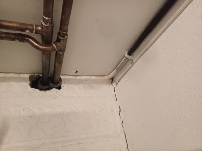 Vägg med kopparrör och kabelkanal. Grov genomföring och sprucken fogning vid golv. Inomhus, närbild.