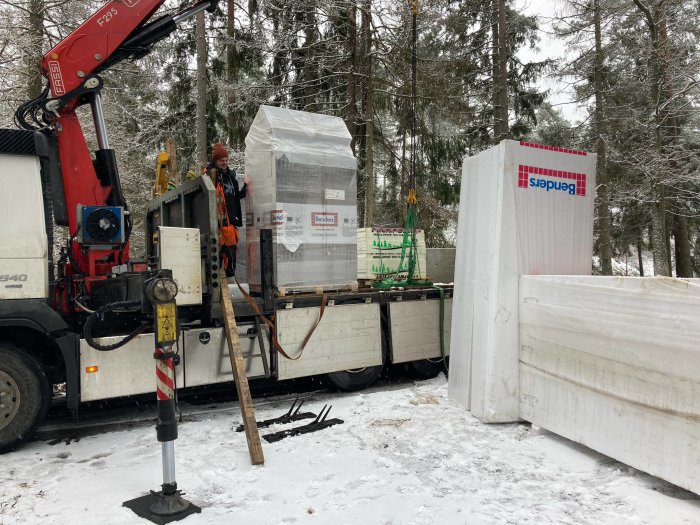 Lastbil med kran lossar byggmaterial i snöigt landskap. Arbetare övervakar. Skog i bakgrunden.