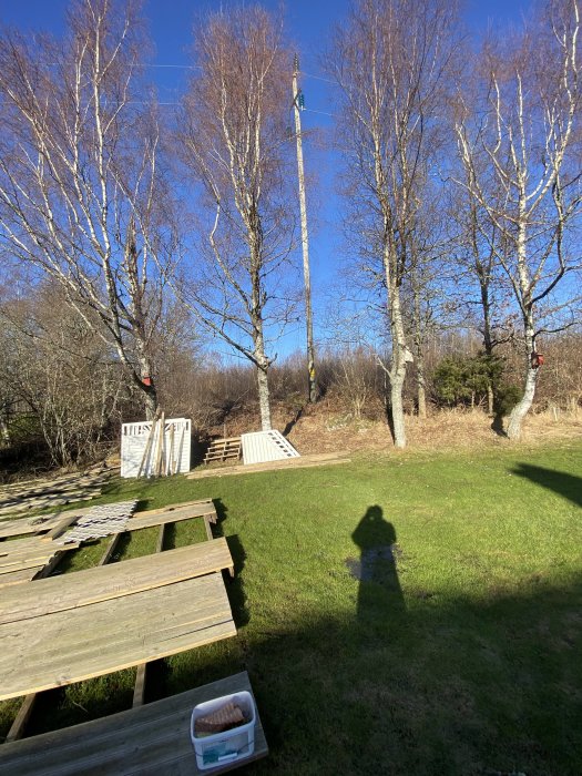 Solig trädgård med träd, gräs, picknickbord, klätterställning, skugga av fotograf, klarblå himmel.