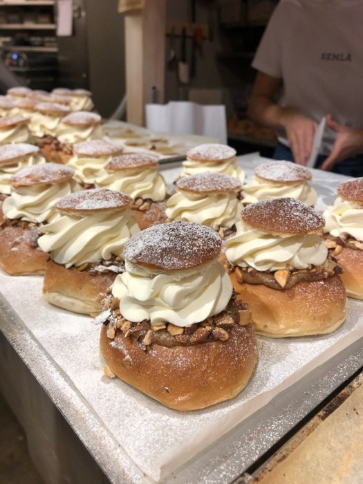 Läckra semlor med grädde och mandelspån, bakverk på hylla i bageri, suddig bakgrund med person.