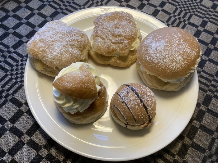 Vitt fat med semlor och bakelser, pudrade med florsocker, på rutig duk.