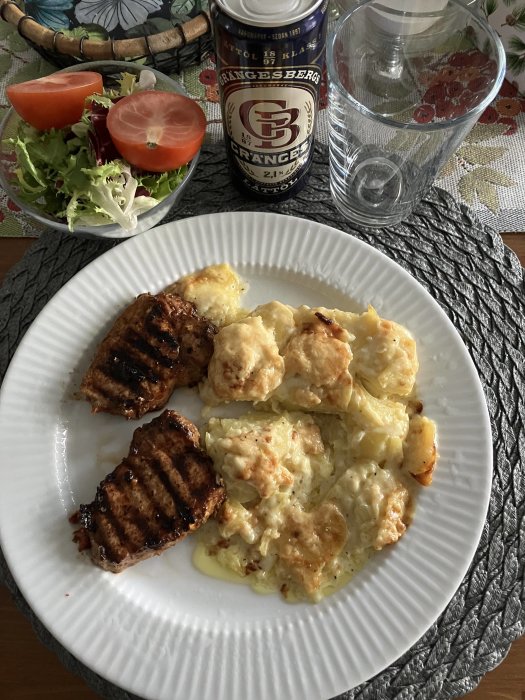 Middagsdukning med grillade köttbitar, gratinerat potatismos, sallad, burköl och ett glas vatten.