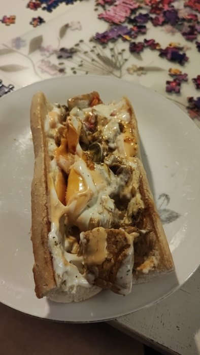 En baguette med fyllning och sås på en tallrik, ovanpå ett bord med pusselbitar i bakgrunden.